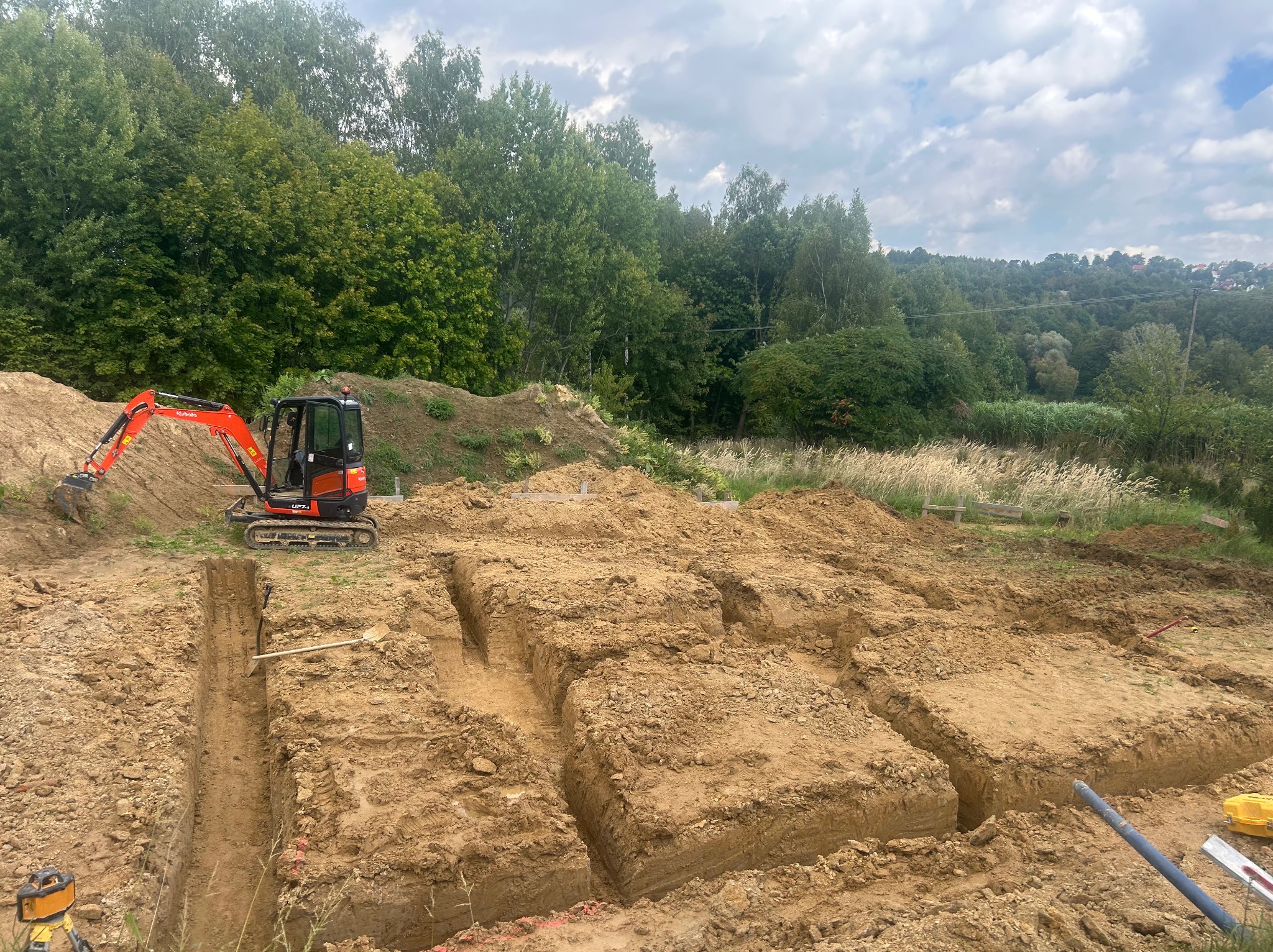Wykopy pod fundamenty z pomarańczową mini koparką Kubota na tle zieleni. Widoczne narzędzia i laserowy niwelator. Prace ziemne w toku, przygotowanie pod budowę.
