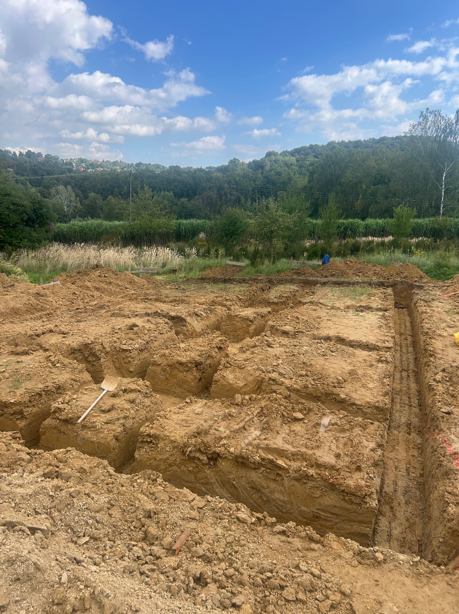 Wykop pod fundamenty w Świątnikach Górnych. Widoczne równe rowy w ziemi, przygotowane pod wylanie fundamentów. Na pierwszym planie łopata wbita w ziemię.