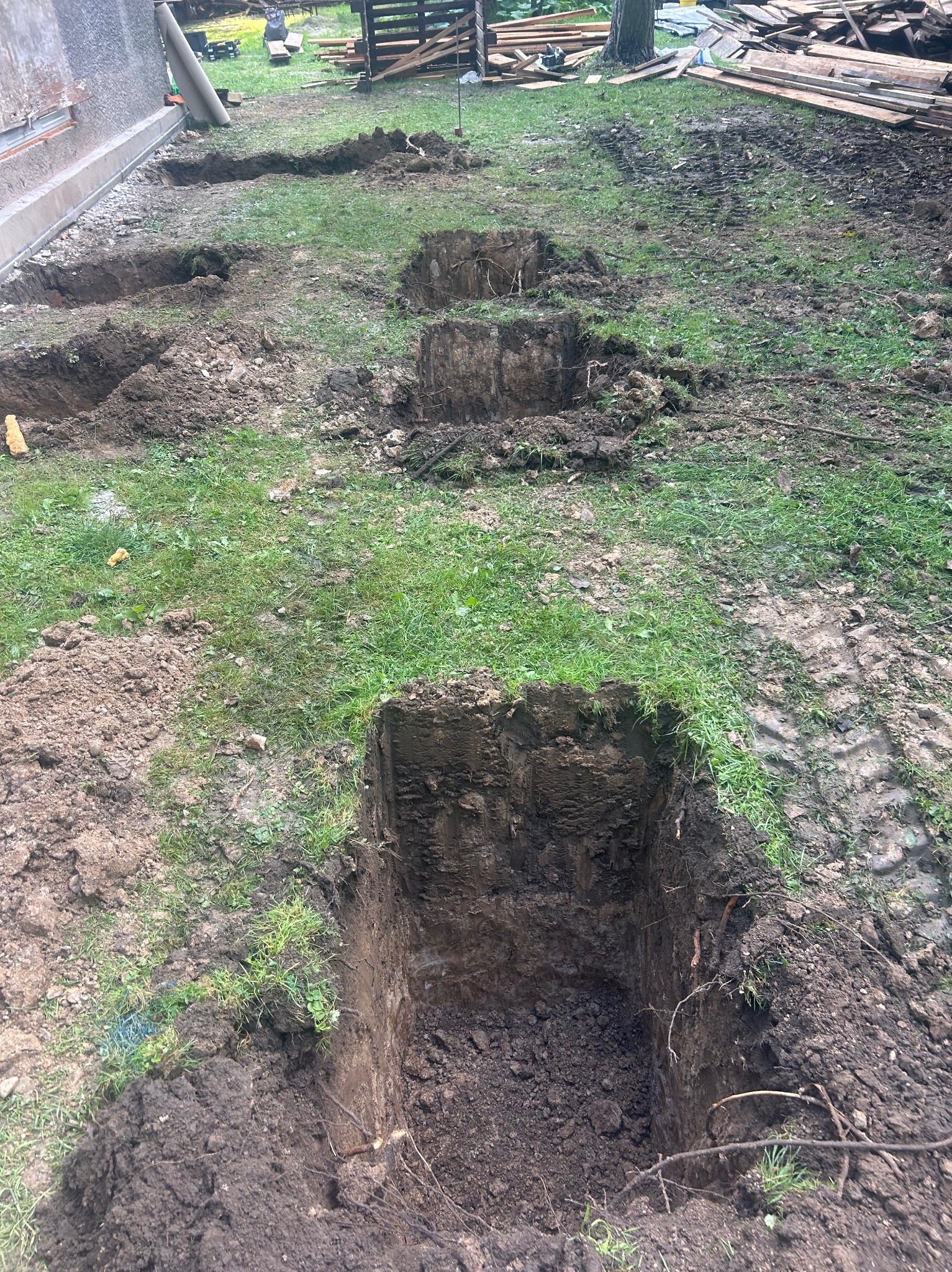 Wykopy pod fundamenty na trawiastym terenie, widoczne ślady opon i składowane drewno w tle. Wyraźne, kwadratowe otwory w ziemi, przygotowane pod budowę.