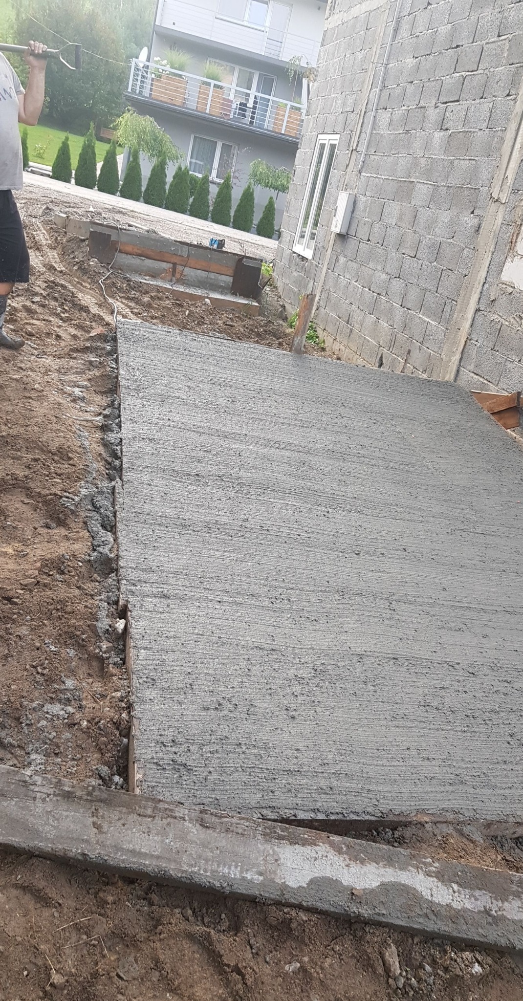 Wylany betonowy podjazd przy domu z bloczków, widoczny pracownik z narzędziem i przygotowane szalunki. Świeży beton o szorstkiej fakturze.