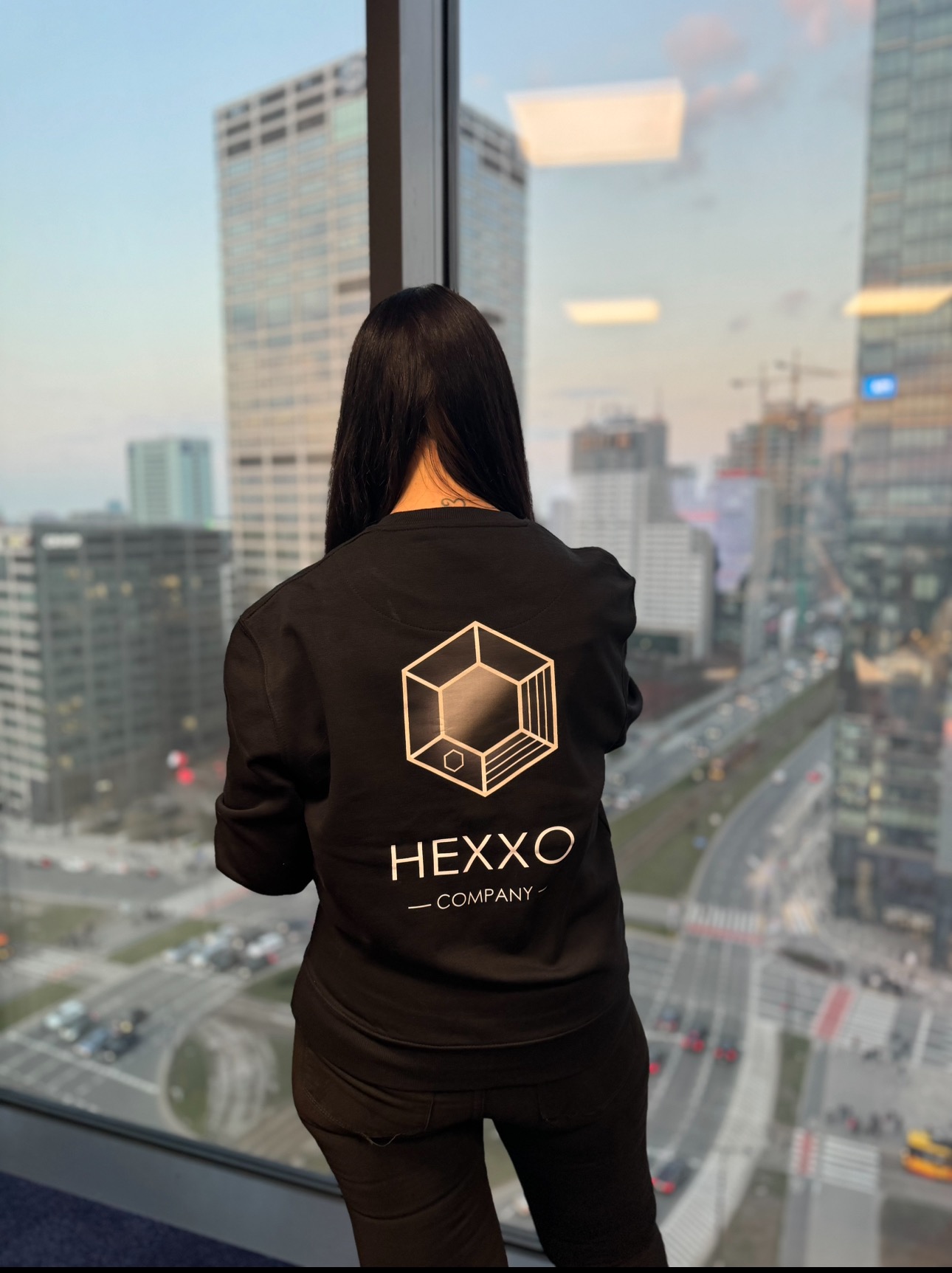 Osoba w bluzie z logo firmy Hexxo patrzy przez okno na panoramę Warszawy. Widok z biura na miasto.