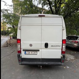 wozimyto - Tył białego busa Fiat Ducato z widoczną tablicą rejestracyjną LSW 45397, zaparkowanego na ulicy w otoczeniu drzew. W tle widoczny budynek.