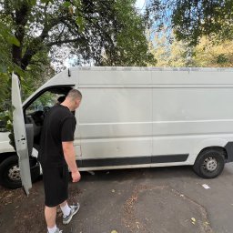 wozimyto - Biały bus dostawczy z otwartymi drzwiami kierowcy, obok stoi mężczyzna w czarnym ubraniu. Widoczne drzewa i fragment chodnika. Bus zaparkowany na ulicy.