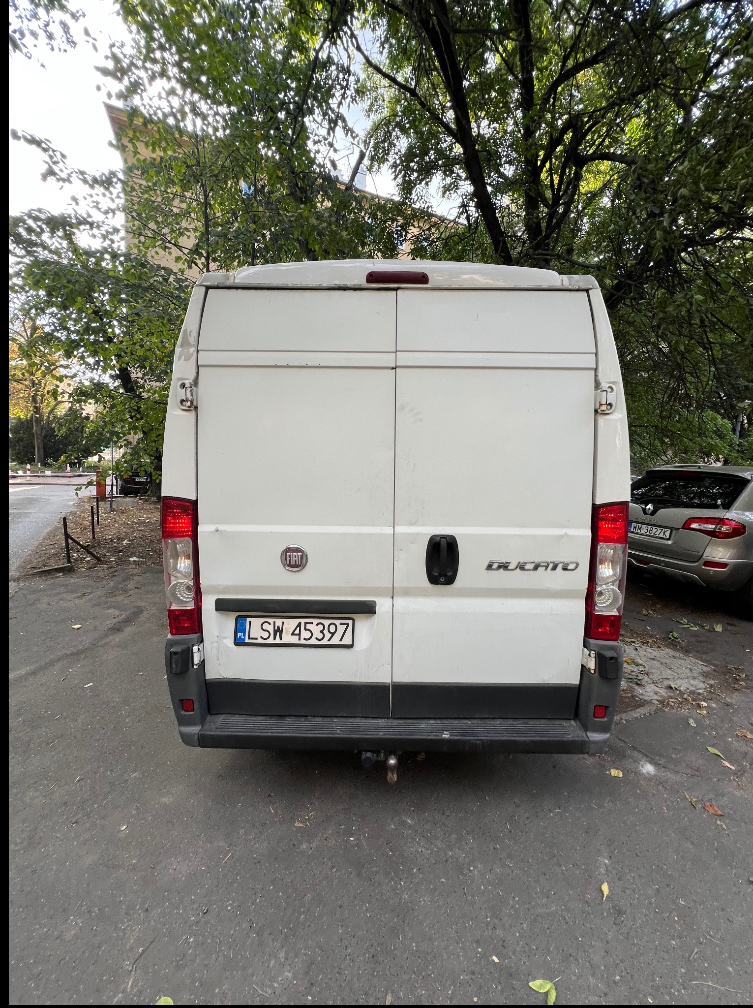 Tył białego busa Fiat Ducato z widoczną tablicą rejestracyjną LSW 45397, zaparkowanego na ulicy w otoczeniu drzew. W tle widoczny budynek.