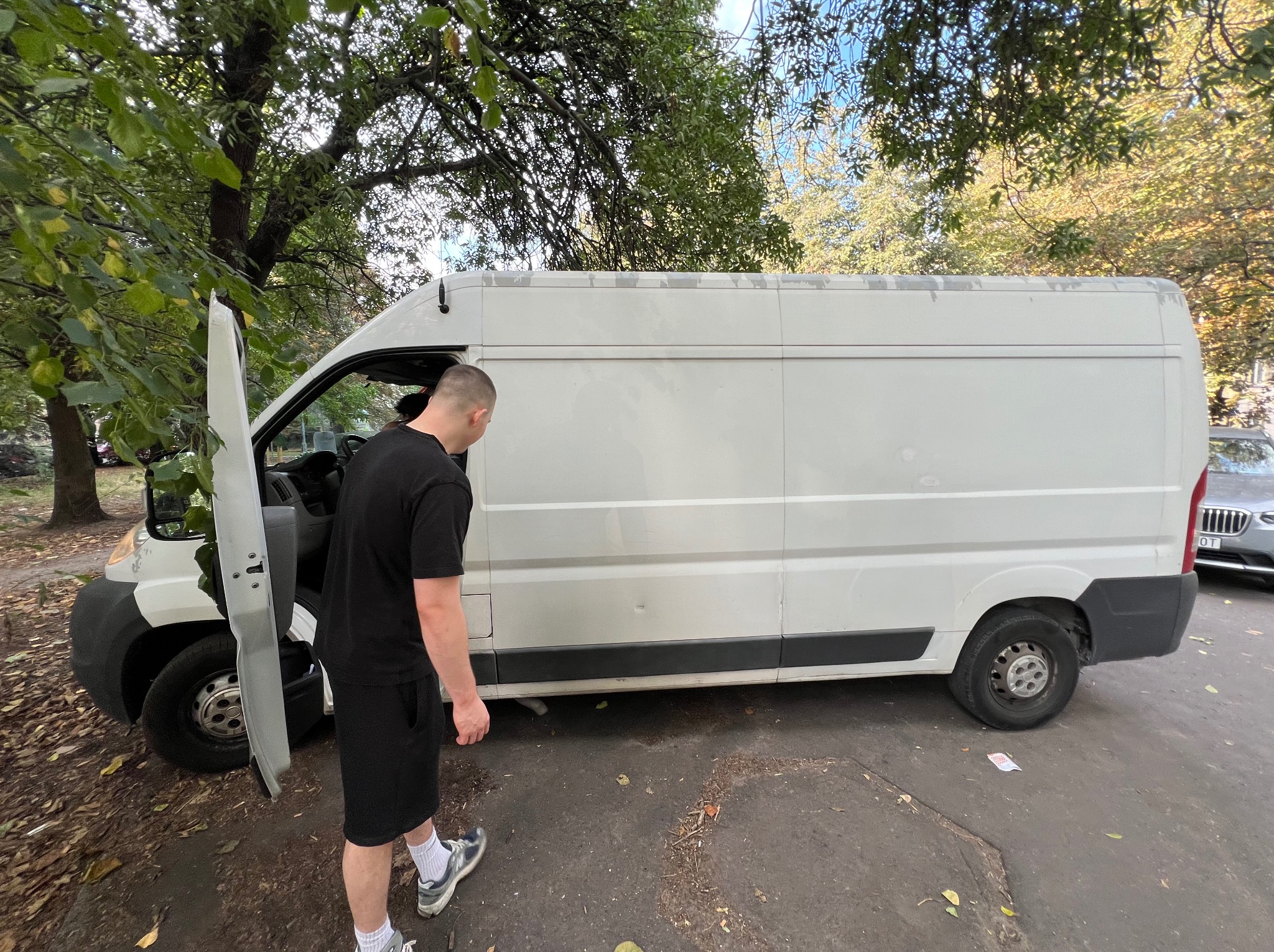 Biały bus dostawczy z otwartymi drzwiami kierowcy, obok stoi mężczyzna w czarnym ubraniu. Widoczne drzewa i fragment chodnika. Bus zaparkowany na ulicy.