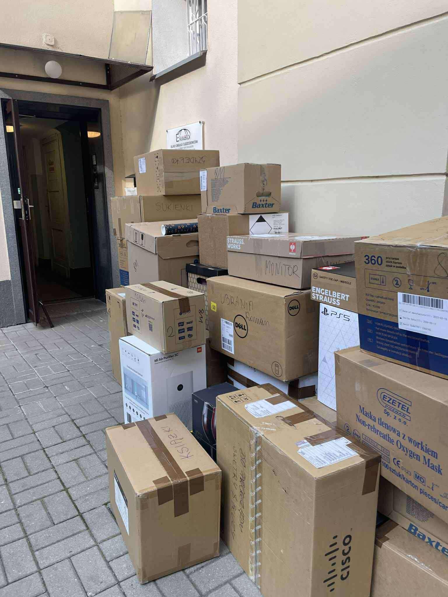Stos kartonowych pudeł przed wejściem do budynku, niektóre z napisem 'Ubrania', 'Książki', 'Sukienki', 'Monitor', 'PS5', sugerujące przeprowadzkę.