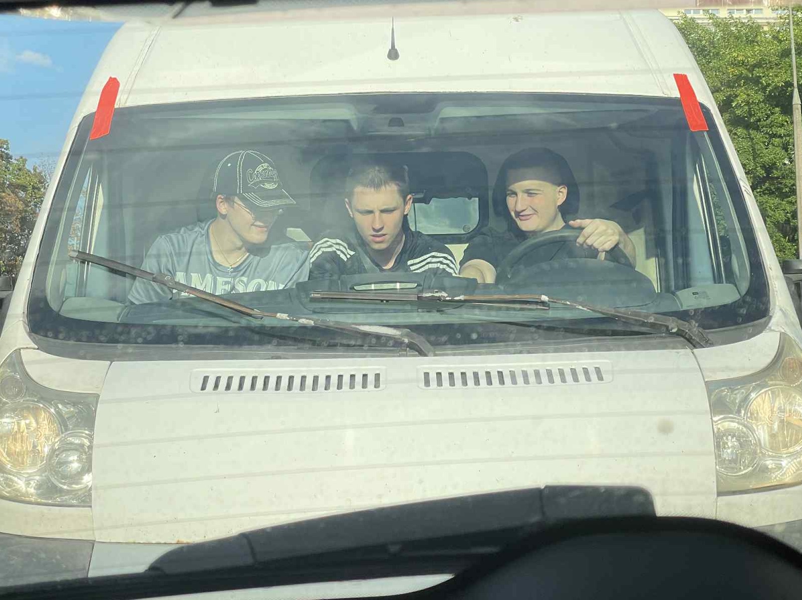 Trzech młodych mężczyzn siedzi w kabinie białego busa, widocznego od przodu, z elementami otoczenia miejskiego w tle.