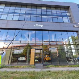 WONDER WASH - Pracownik w kamizelce Wonder Wash myje okna na wysokości w salonie Jeep, używając długiej lancy. Widoczne odbicia w oknach i otoczenie budynku.