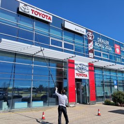 WONDER WASH - Mycie okien na wysokości w salonie Toyota Auto Park. Pracownik używa teleskopowego kija do czyszczenia przeszklonej fasady budynku dealera samochodowego.