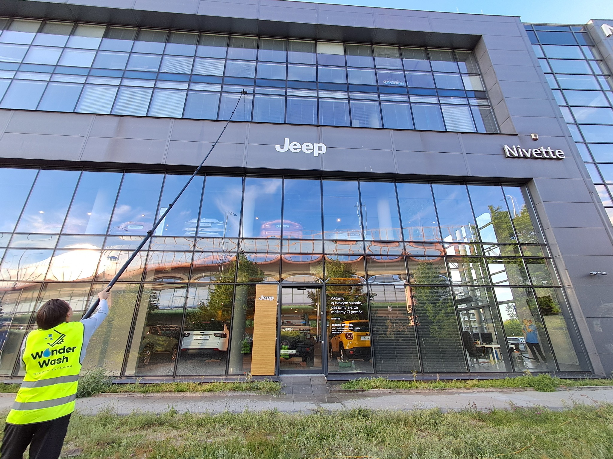 Pracownik w kamizelce Wonder Wash myje okna na wysokości w salonie Jeep, używając długiej lancy. Widoczne odbicia w oknach i otoczenie budynku.