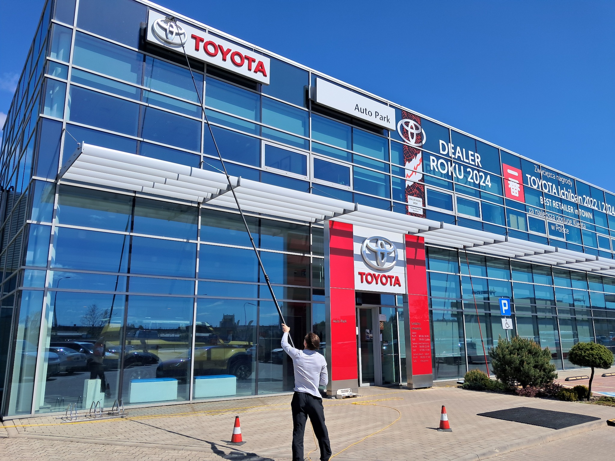 Mycie okien na wysokości w salonie Toyota Auto Park. Pracownik używa teleskopowego kija do czyszczenia przeszklonej fasady budynku dealera samochodowego.