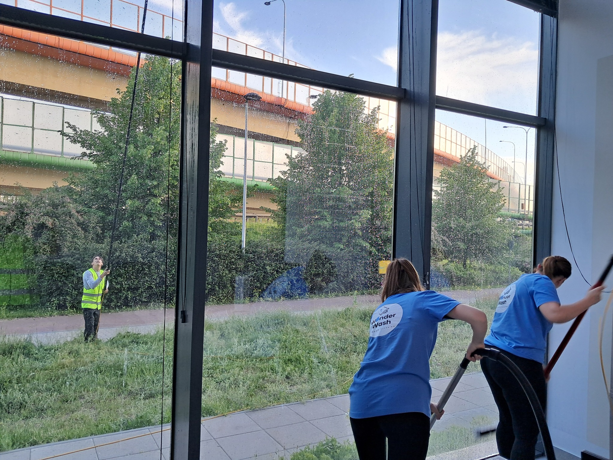 Mycie okien w biurowcu: dwie pracownice w błękitnych koszulkach czyszczą szyby od wewnątrz, trzeci pracownik spryskuje okna z zewnątrz, widok na zieleń i ulicę.