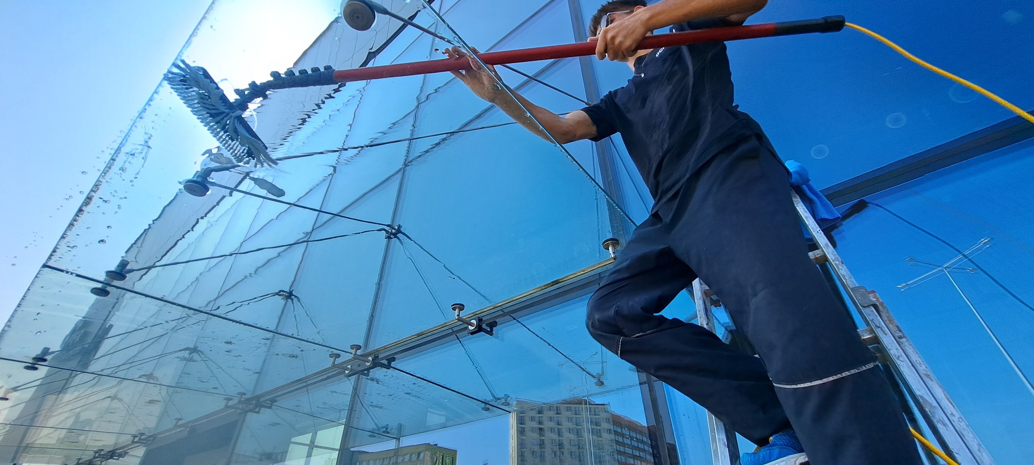 Pracownik na drabinie myje szklaną fasadę budynku przy użyciu szczotki na teleskopowym kiju. Widoczne krople wody i odbicie budynków w szybach.