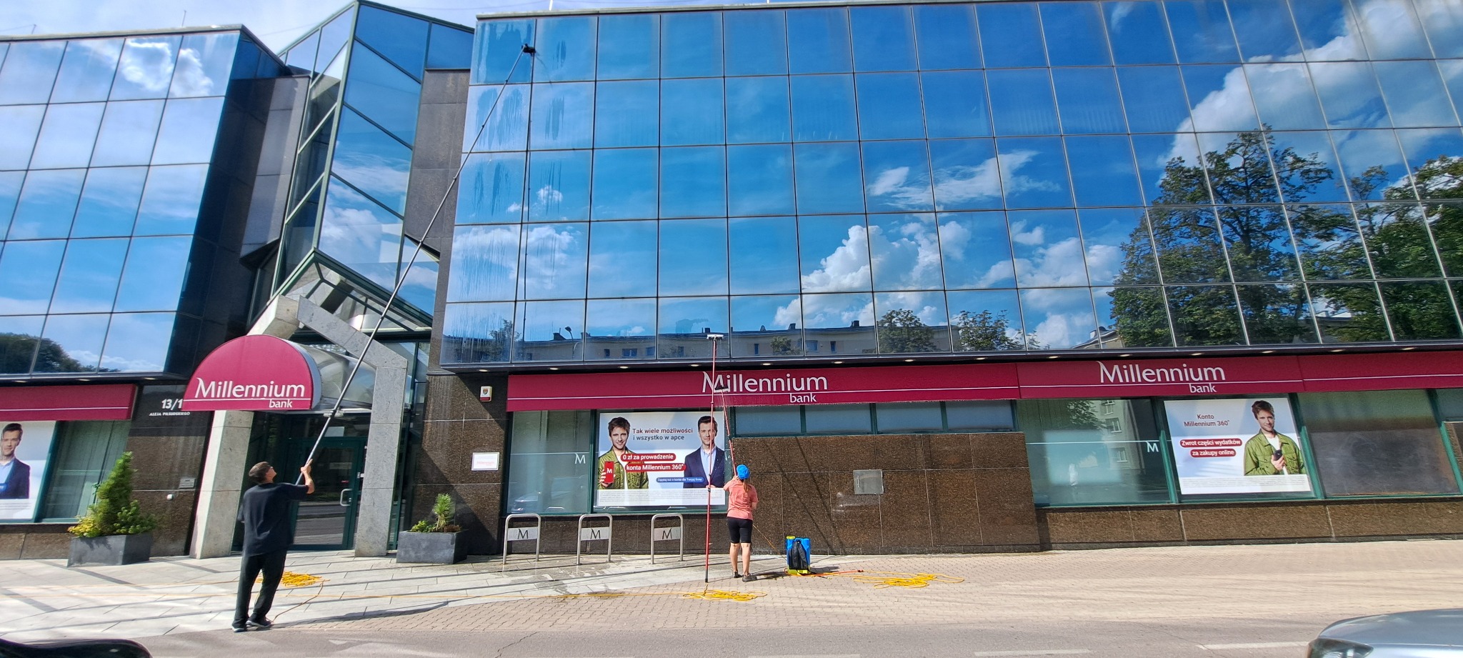 Mycie okien biurowca Millennium Bank z użyciem tyczek teleskopowych. W tle odbicie nieba i drzew w szklanej fasadzie, dwóch pracowników w trakcie pracy.