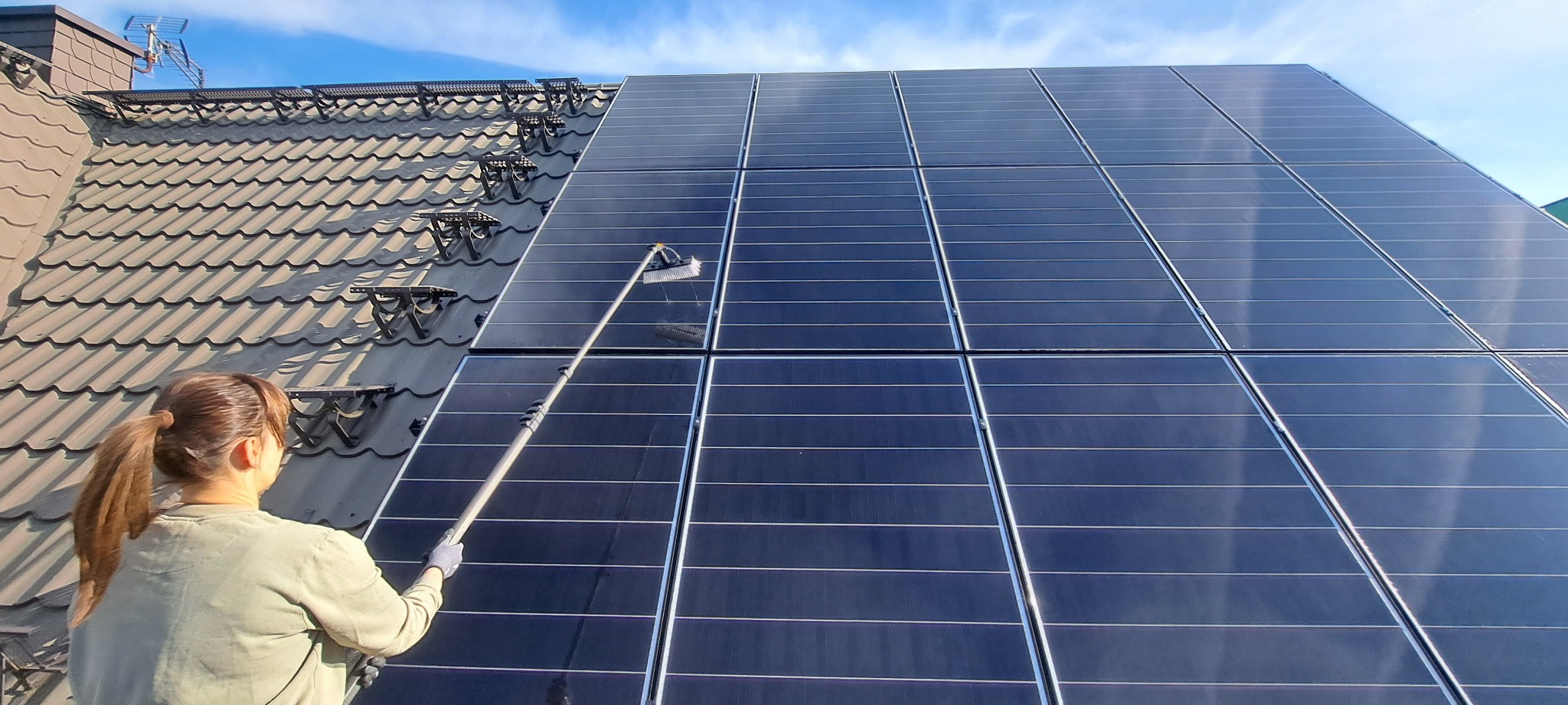 Kobieta czyści panele słoneczne na dachu domu za pomocą szczotki na długim teleskopowym kiju. Widoczne dachówki i błękitne niebo.