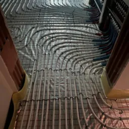 Białe rurki ogrzewania podłogowego ułożone na folii z siatką, widok z góry, etap prac instalacyjnych przed wylaniem posadzki. Rozdzielacz z rurami w tle.