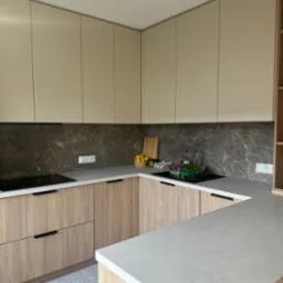 Nowoczesna kuchnia w beżach i szarościach. Jasne, drewnopodobne fronty szafek dolnych, gładkie, beżowe szafki górne. Szary blat i marmurowa ściana. Geometryczna podłoga.