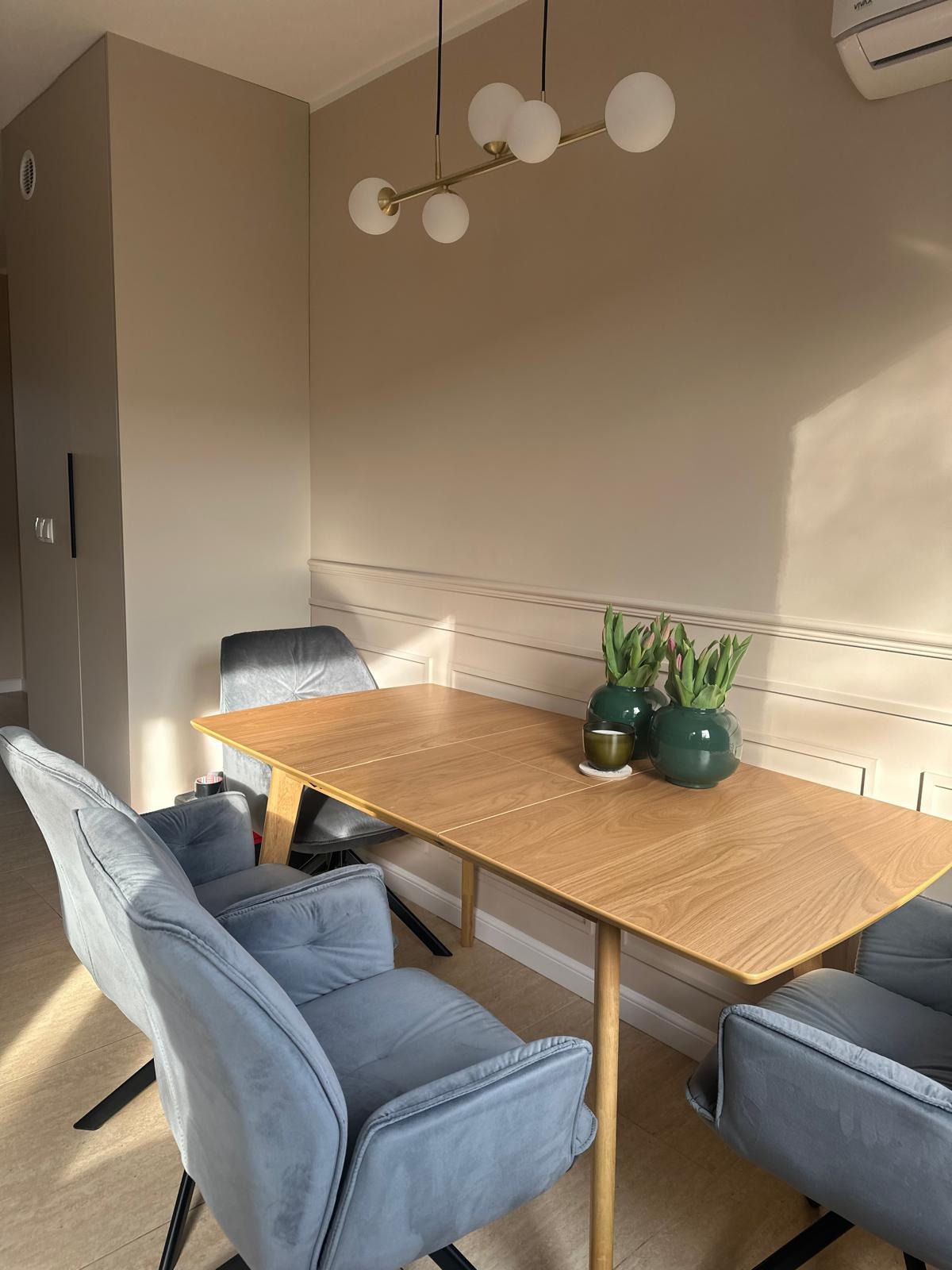 Elegancka jadalnia w beżach: drewniany stół z tulipanami w wazonach, welurowe krzesła w odcieniach szarości, dekoracyjna listwa ścienna i nowoczesna lampa.