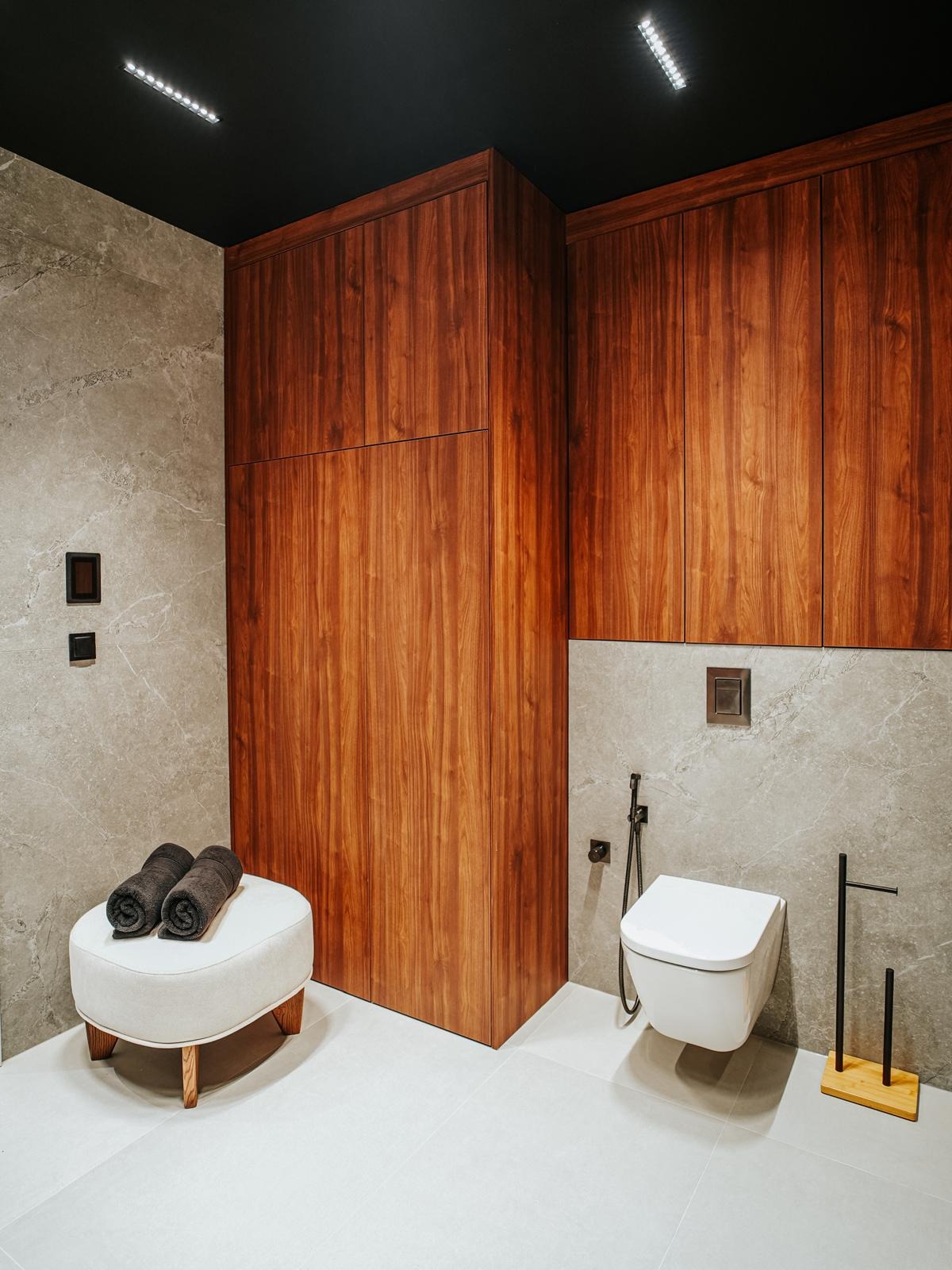Nowoczesna łazienka z zabudową z drewna orzechowego, szarą płytką imitującą marmur i podwieszaną toaletą. Oświetlenie LED w czarnym suficie.