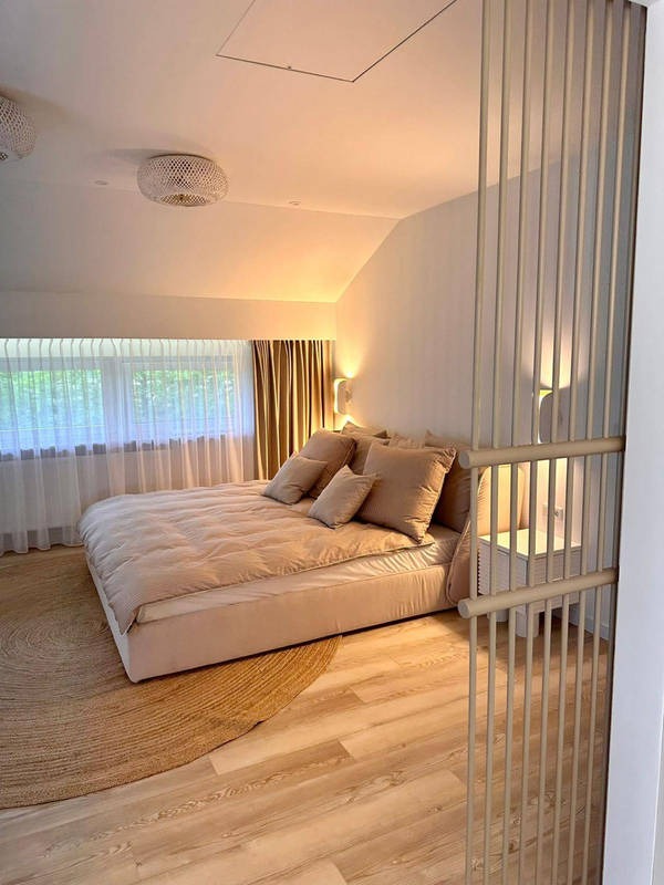 Beżowa sypialnia na poddaszu z drewnianą podłogą i okrągłym dywanem. Widoczny ażurowy przepierzenie i łóżko z beżową pościelą. Okno z zasłonami w tle.