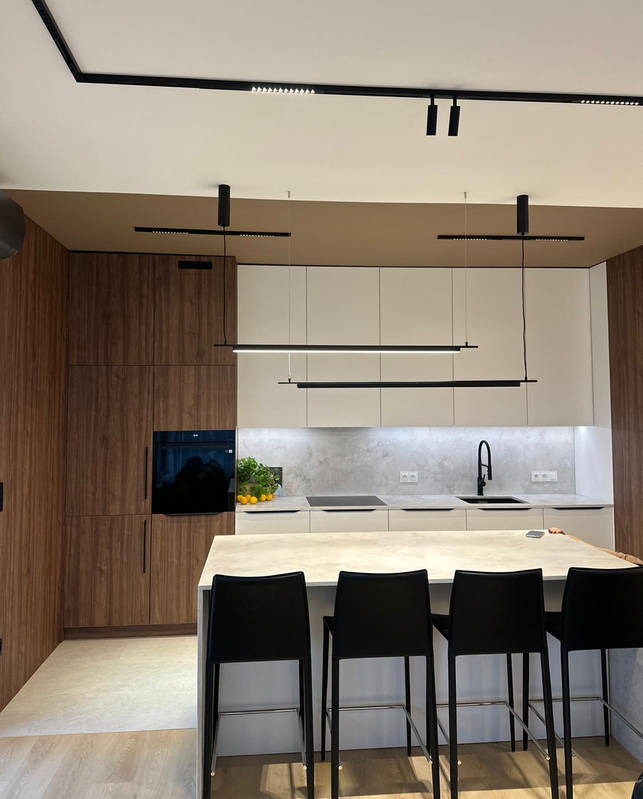 Nowoczesna kuchnia po remoncie: drewniane szafki z piekarnikiem, białe szafki wiszące, wyspa z czarnymi hokerami i designerskie, podłużne lampy sufitowe.