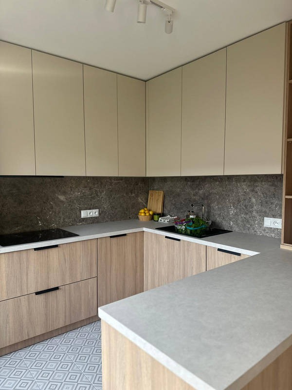 Jasna, narożna kuchnia z drewnianymi frontami szafek dolnych i beżowymi górnymi, szary blat i marmurowy backsplash, wzorzysta podłoga. Nowoczesny design.