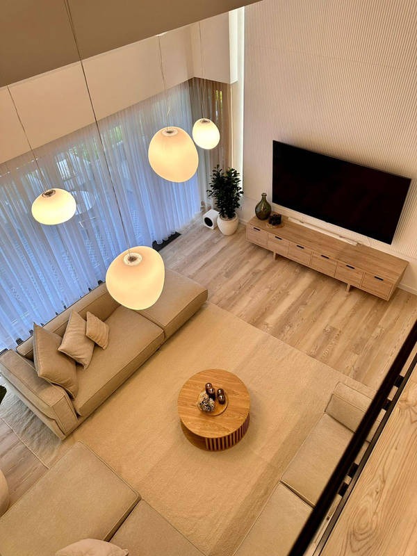 Nowoczesny salon z wysokim sufitem, beżową sofą, drewnianym stolikiem kawowym i podłogą z paneli. Cztery okrągłe lampy zwisają z sufitu.