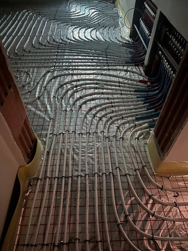 Białe rurki ogrzewania podłogowego ułożone na folii z siatką, widok z góry, etap prac instalacyjnych przed wylaniem posadzki. Rozdzielacz z rurami w tle.