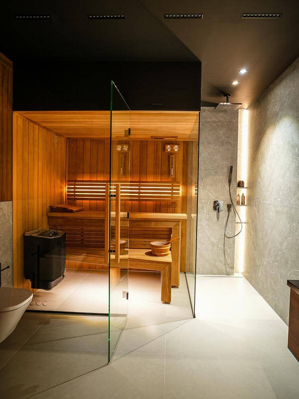 Nowoczesna łazienka z sauną: drewniane wnętrze sauny za szklaną ścianą, marmurowe płytki, oświetlenie punktowe i minimalistyczny design.