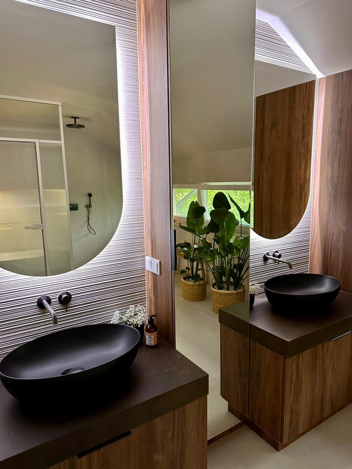 Nowoczesna łazienka z dwoma umywalkami w kolorze czarnym, drewnianymi szafkami i podświetlanymi lustrami owalnymi. W tle rośliny doniczkowe i kabina prysznicowa.