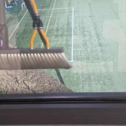 Perfect Shine Firma sprzątająca Mateusz Fąfara - Szczotka do mycia okien na długim kiju, czyszcząca szybę z widokiem na boisko. Widoczne smugi i zabrudzenia na szybie. Czarna rama okienna w dolnej części kadru.