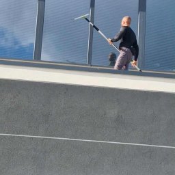 Perfect Shine Firma sprzątająca Mateusz Fąfara - Pracownik w trakcie mycia okien na wysokości przy użyciu specjalistycznego sprzętu. Widoczne odbicie nieba w oknach. Szary budynek w tle.