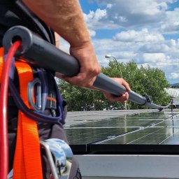 Perfect Shine Firma sprzątająca Mateusz Fąfara - Mycie paneli słonecznych na dachu w Rzeszowie. Widoczny fragment uprzęży i teleskopowa szczotka do czyszczenia. Błękitne niebo w tle.