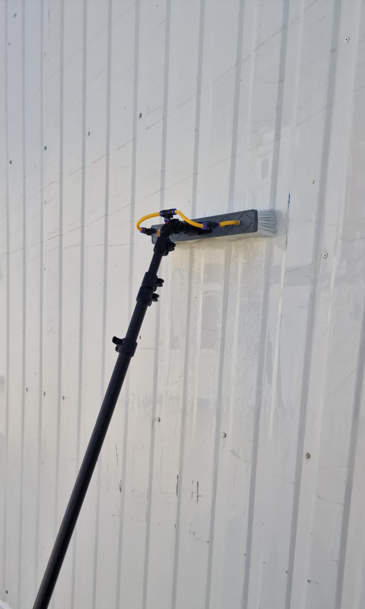 Mycie elewacji przemysłowej lancą teleskopową z szczotką. Widoczne ślady wody i czyszczona powierzchnia. Detal narzędzia pracy.