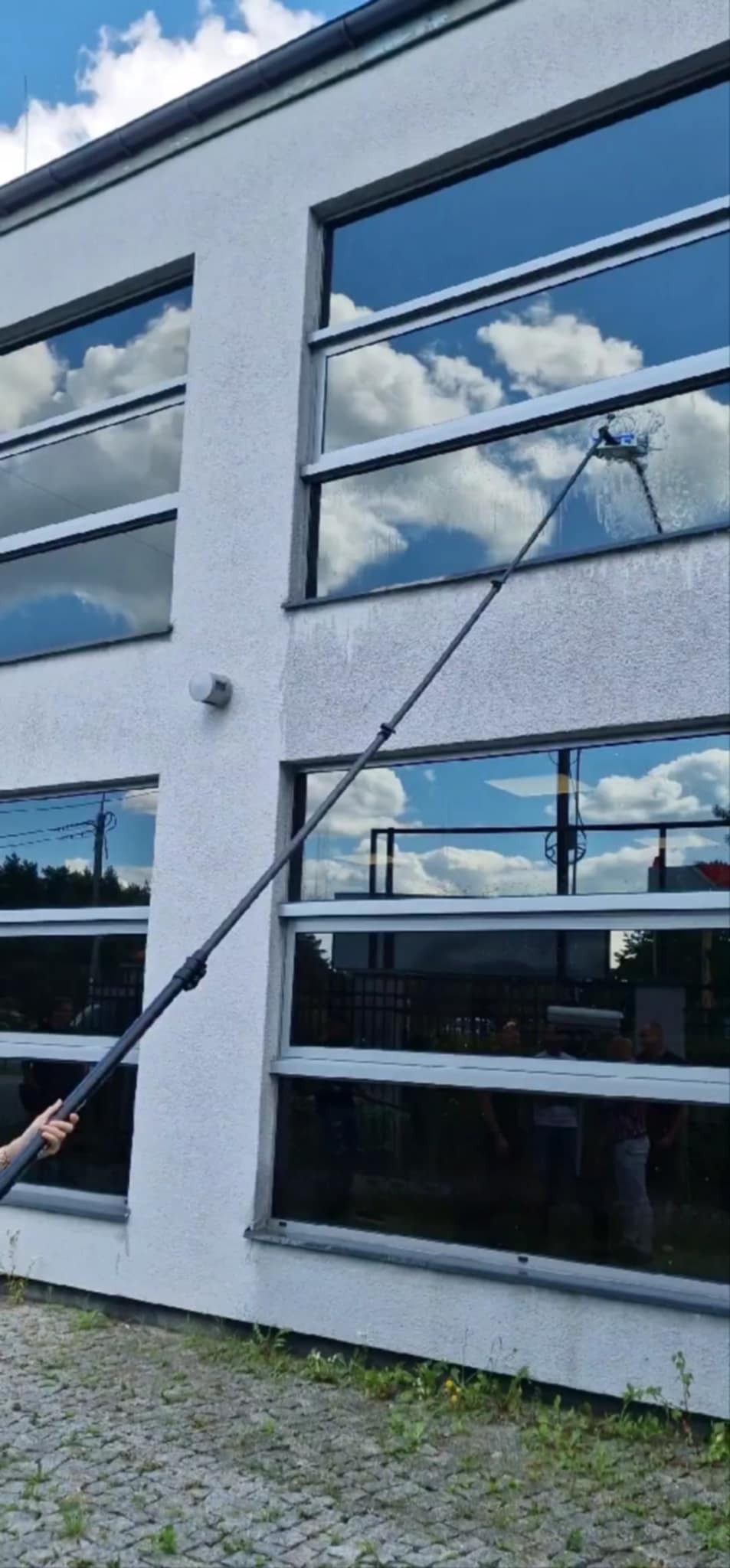 Mycie okien na wysokości z użyciem teleskopowego kija. Na oknach odbija się błękitne niebo z chmurami. Widoczna elewacja budynku i fragment chodnika.