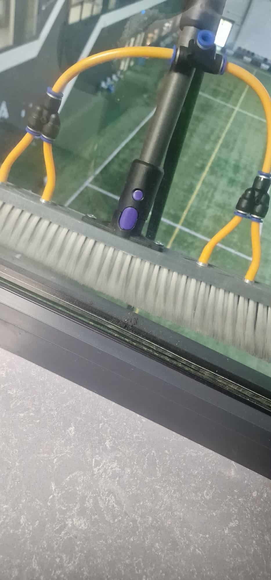 Szczotka teleskopowa z żółtymi wężami myje brudne okno. Widoczne zacieki i kurz na ramie okiennej. W tle zielona przestrzeń.