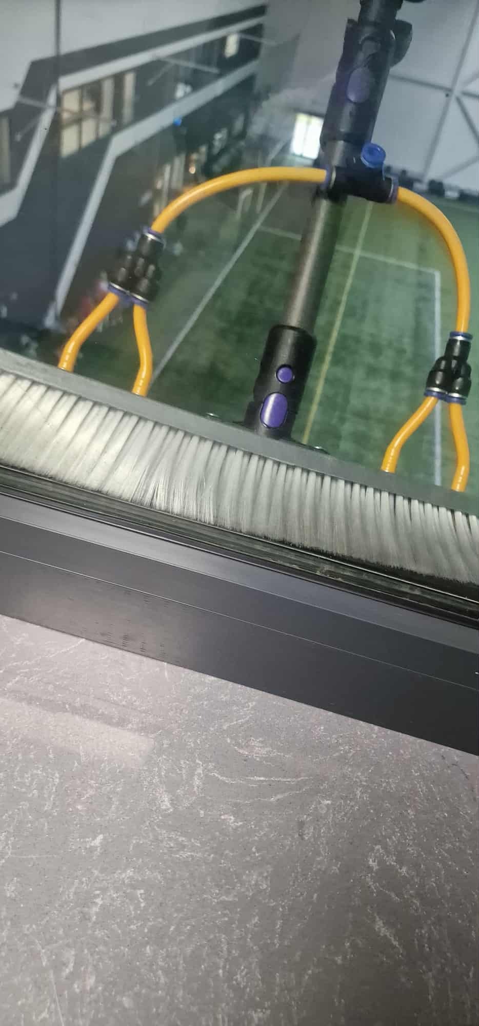Szczotka do mycia okien na wysięgniku, z żółtymi przewodami doprowadzającymi wodę, oparta o szybę z widokiem na halę sportową. Detal narzędzia.