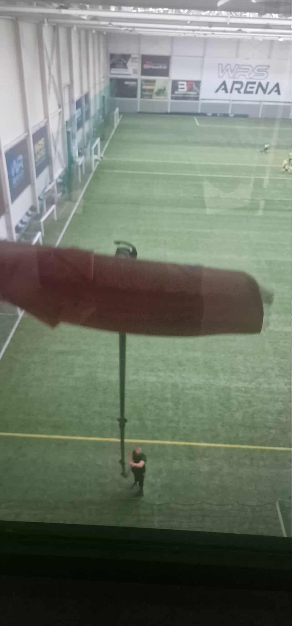 Wnętrze hali sportowej z zieloną nawierzchnią, widoczne osoby i sprzęt do czyszczenia. Ujęcie z góry, z widocznym fragmentem ekranu.