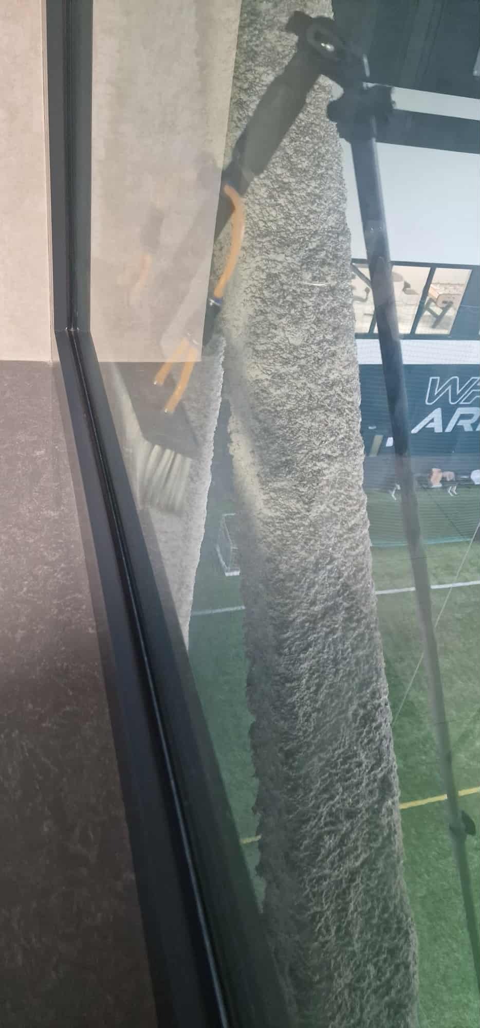 Mycie okna z zewnątrz za pomocą szczotki na teleskopowym kiju. Widoczny fragment elewacji budynku i boisko sportowe za oknem. Praca na wysokości.