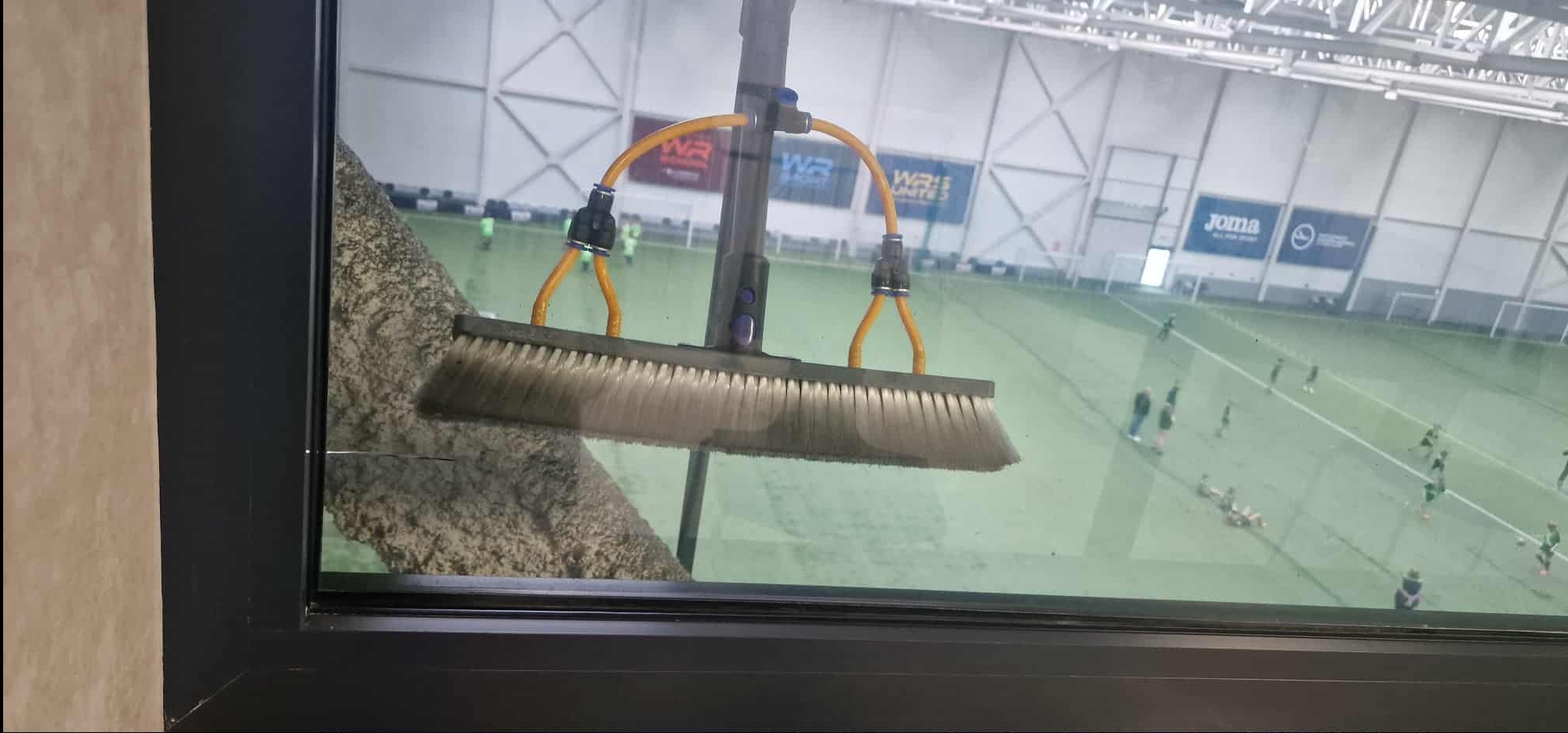 Mycie okien w hali sportowej z użyciem specjalistycznego sprzętu z szczotką i dopływem wody. Widoczny fragment okna i grający w tle piłkarze.