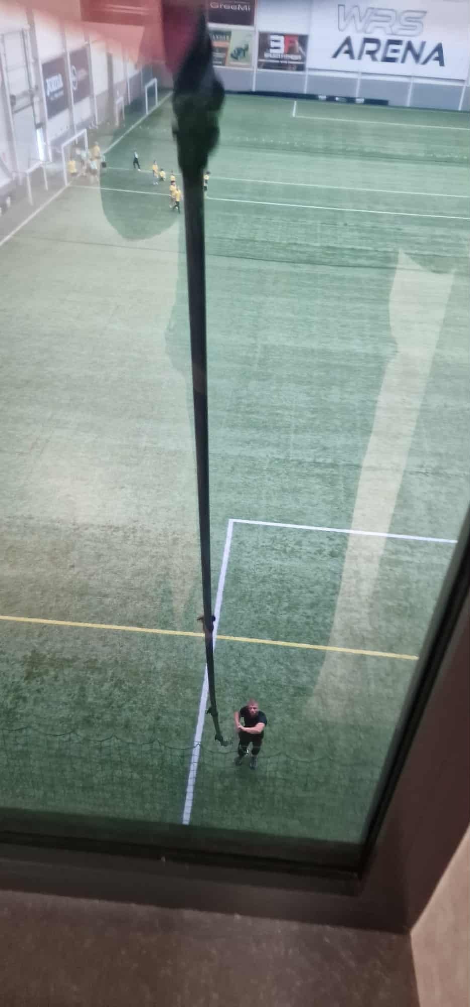 Widok z okna na halę sportową z zieloną murawą, gdzie odbywa się mecz piłki nożnej. Na pierwszym planie widoczna rama okienna i fragment masztu flagowego.