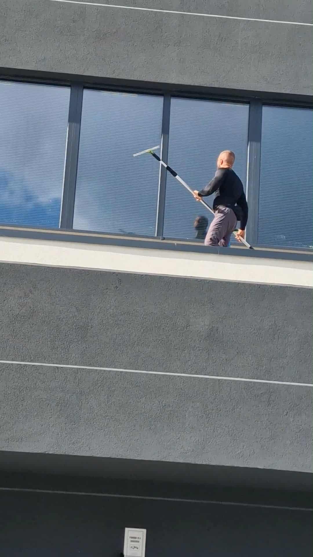 Mężczyzna czyści okna na wysokości za pomocą długiej myjki. Widoczne odbicie w oknach i szara elewacja budynku z poziomymi liniami.
