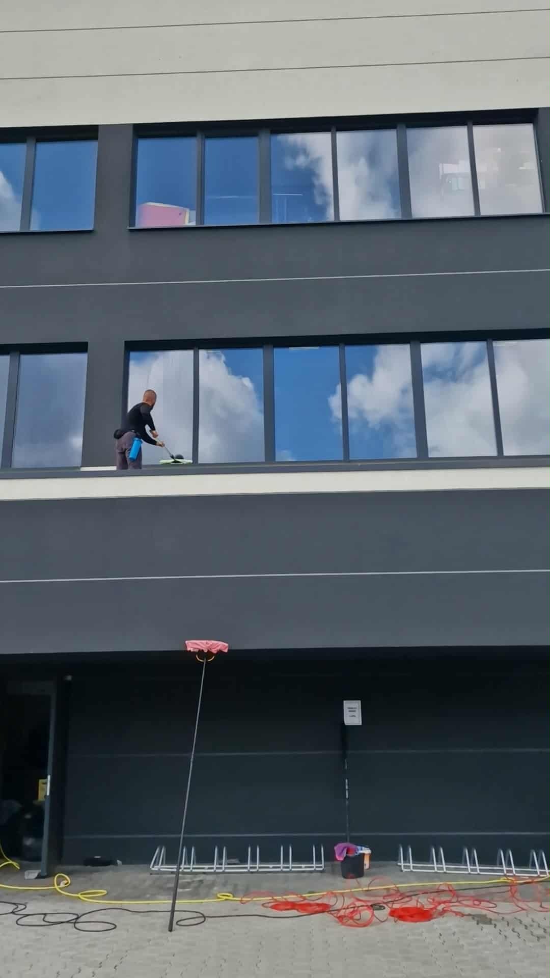 Pracownik myje okna na wysokości w biurowcu, używając specjalistycznego sprzętu. W oknach odbija się błękitne niebo z chmurami. Na dole widoczne narzędzia pracy.