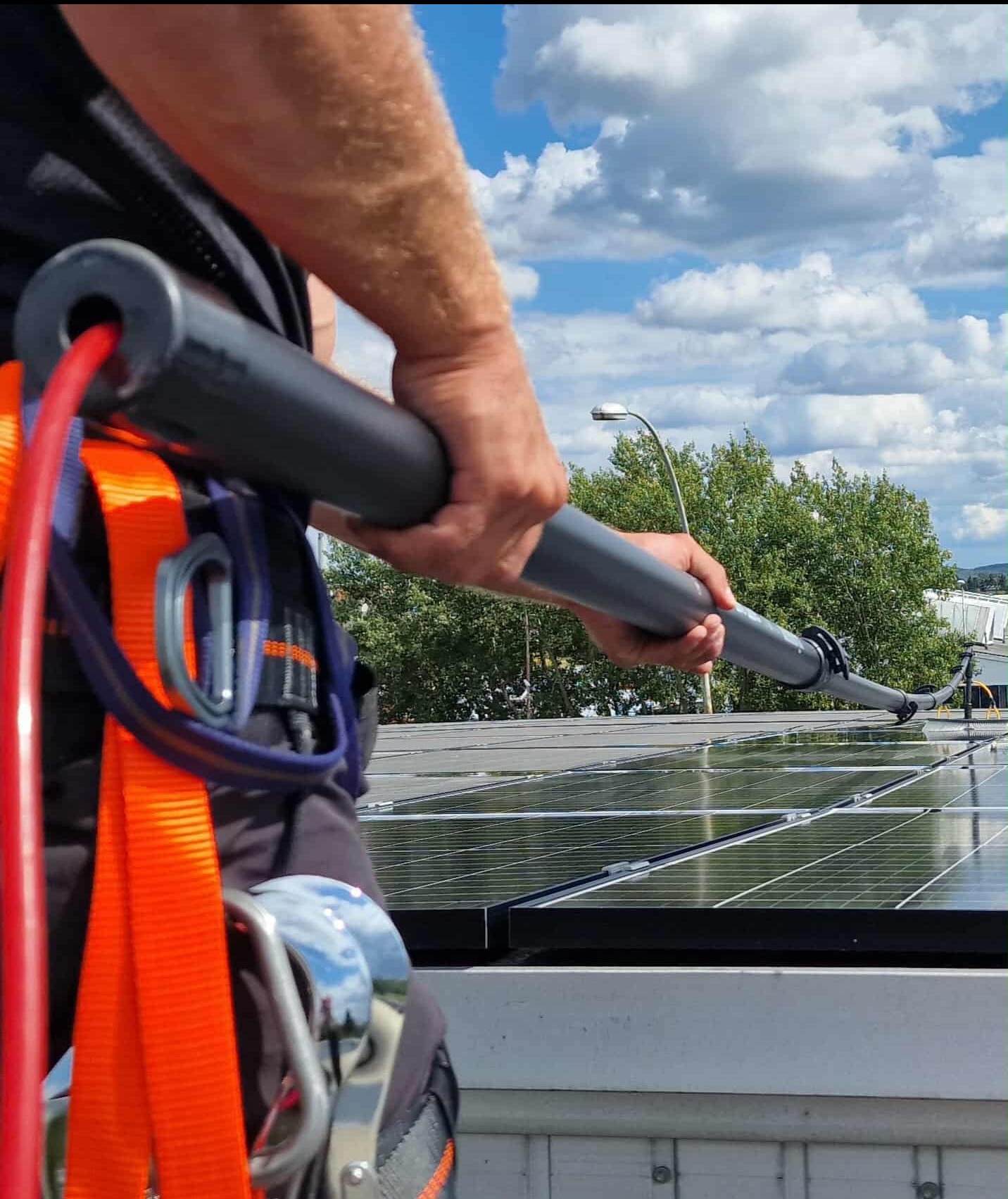 Mycie paneli słonecznych na dachu w Rzeszowie. Widoczny fragment uprzęży i teleskopowa szczotka do czyszczenia. Błękitne niebo w tle.