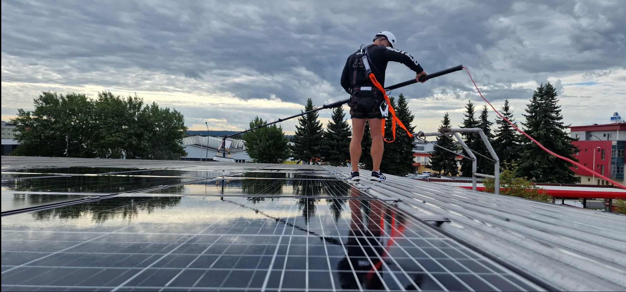 Pracownik w kasku i uprzęży myje panele słoneczne na dachu budynku za pomocą długiej szczotki. W tle pochmurne niebo i drzewa.