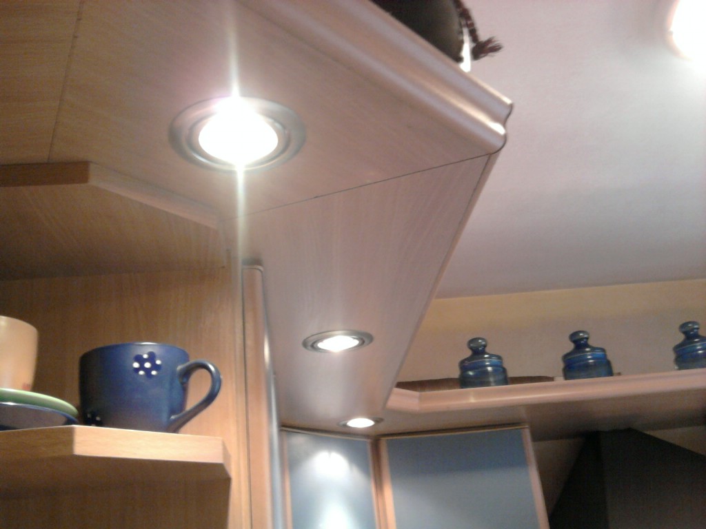 Fragment zabudowy kuchennej z oświetleniem punktowym LED, widoczne szafki z jasnego drewna i niebieskie fronty, na półce dekoracyjne słoiczki oraz niebieski kubek w białe kropki.