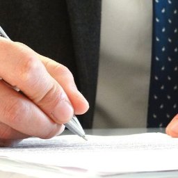 KANCELARIA ADWOKACKA ADWOKAT PRZEMYSŁAW WOJTACHA - Ręka w garniturze pisze na papierze srebrnym długopisem. Mężczyzna w trakcie sporządzania dokumentu. Widoczny fragment krawata w granatowe wzory.