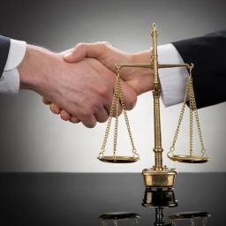 KANCELARIA ADWOKACKA ADWOKAT PRZEMYSŁAW WOJTACHA - Uścisk dłoni i złote wagi Temidy na czarnym blacie, szare tło. Symbol zgody prawnej i sprawiedliwości w biznesie. Detal dłoni i wagi.