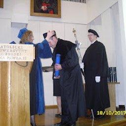 KANCELARIA ADWOKACKA ADWOKAT PRZEMYSŁAW WOJTACHA - Wręczenie dyplomu absolwentowi KUL podczas uroczystości. Absolwent w czarnej todze, odbiera gratulacje od osoby wręczającej dyplom. W tle duchowny.