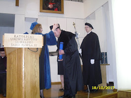 Wręczenie dyplomu absolwentowi KUL podczas uroczystości. Absolwent w czarnej todze, odbiera gratulacje od osoby wręczającej dyplom. W tle duchowny.